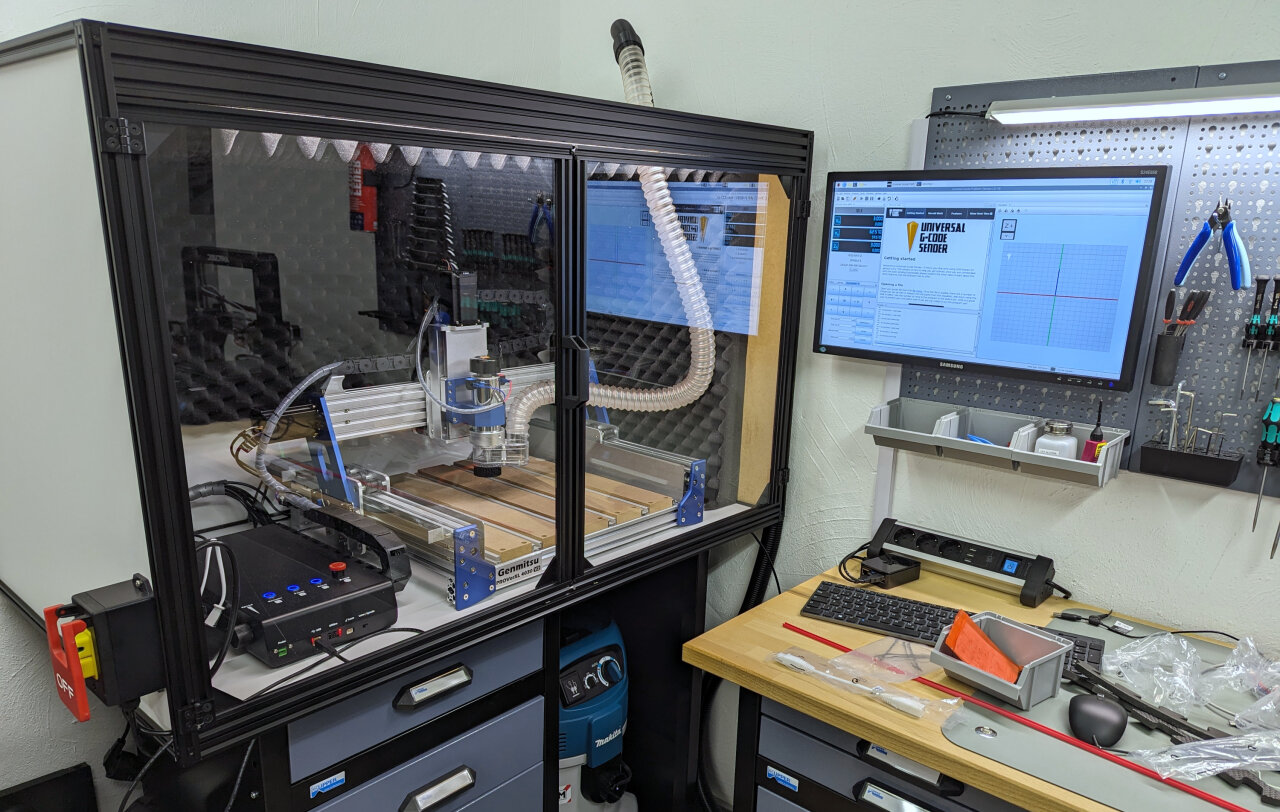 Photo of my setup. On a workbench on the left sits an enclosure made out of aluminium extrusions and MDF sheets at the sides. The front is acrylic glass and through it the CNC machine and its dust extraction hose are visible. To the right is another workbench with tool holders and a screen through which the CNC can be controlled.